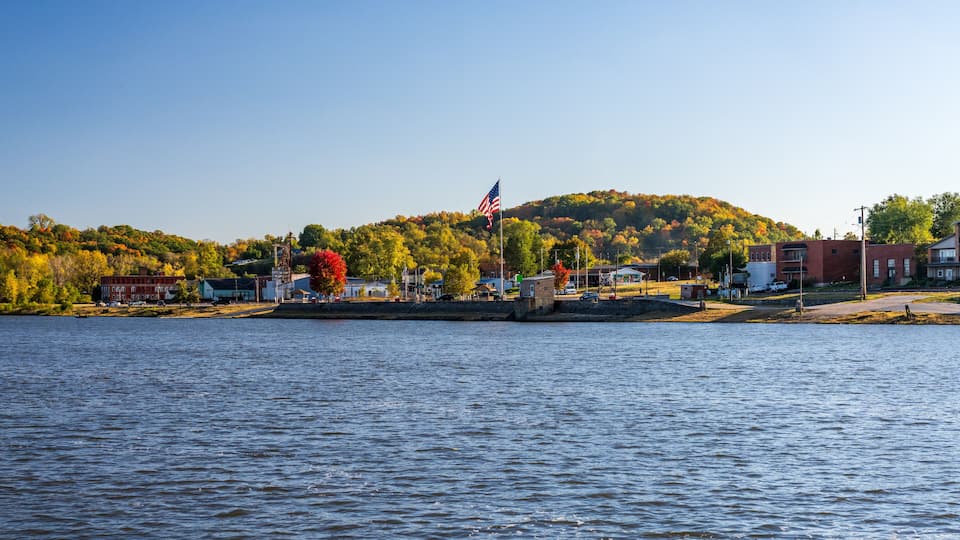 A waterfront town with a hill in the background, a flag on a pole, and buildings along the shore.
