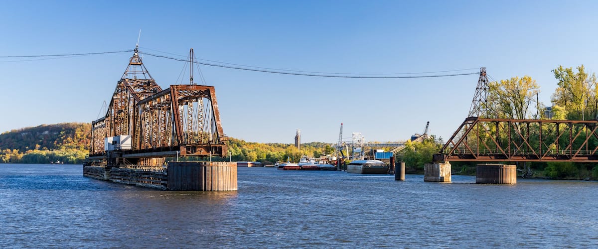 A river with a large, rusted metal bridge and a smaller bridge in the background.