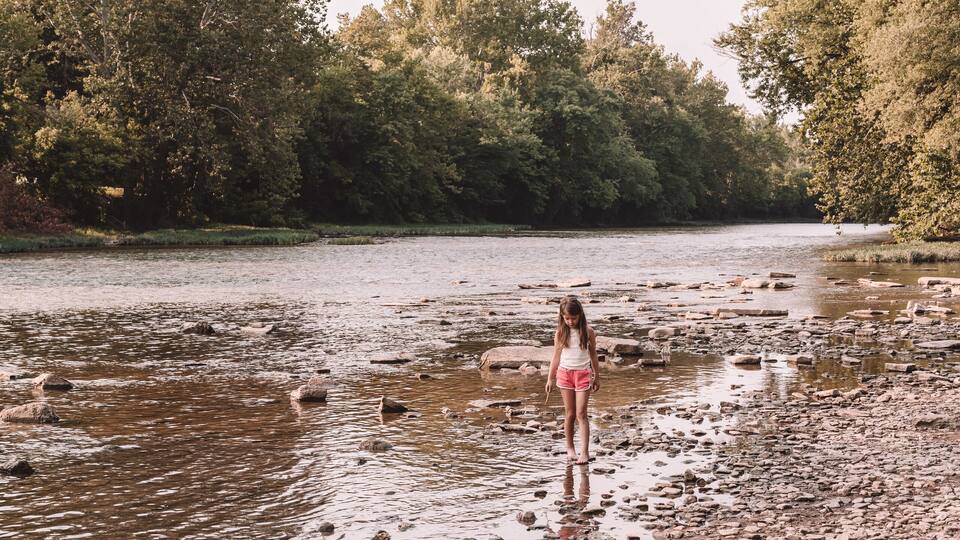 A riverbank with a kayak, paddle, and gear, and a person standing in the water.
