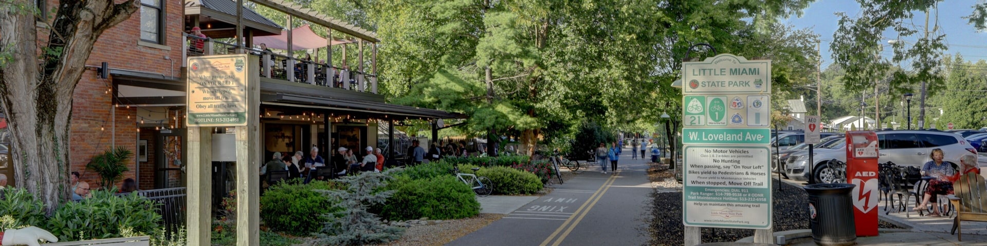 View of a brick building with a restaurant and people dining outside under the trees, beside a pathway for pedestrians, Loveland, Ohio, United States.