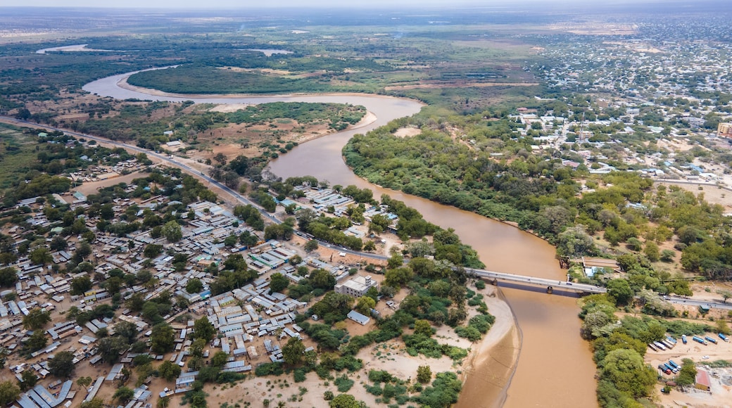 Aerial view of the brown Tana River snaking through the arid landscape, contrasting with the dense green vegetation and buildings, Garissa, Kenya.