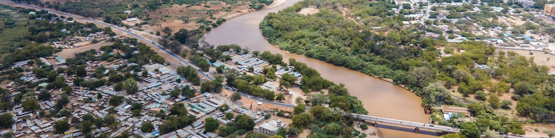 Aerial view of the brown Tana River snaking through the arid landscape, contrasting with the dense green vegetation and buildings, Garissa, Kenya.