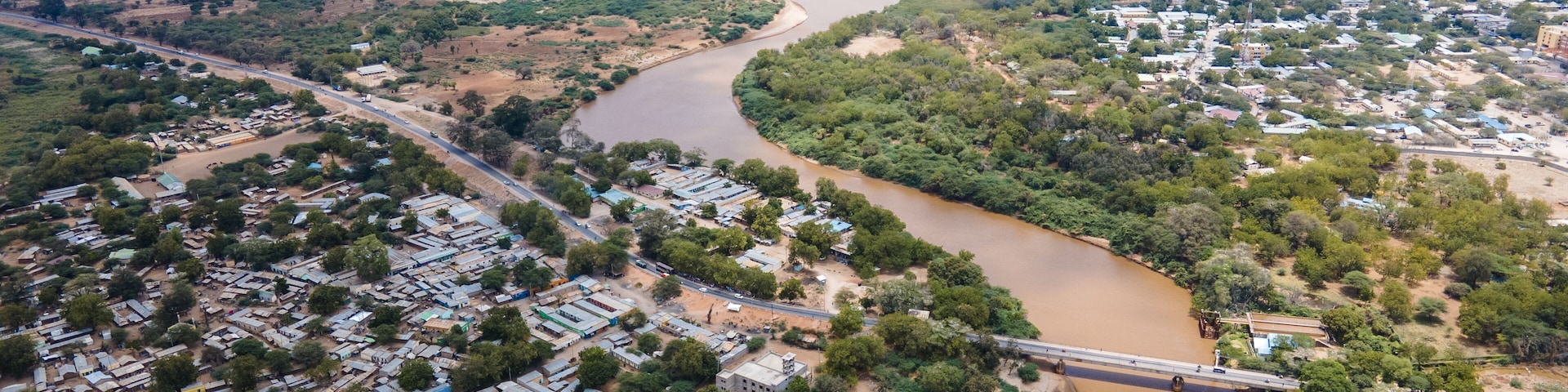 Aerial view of the brown Tana River snaking through the arid landscape, contrasting with the dense green vegetation and buildings, Garissa, Kenya.