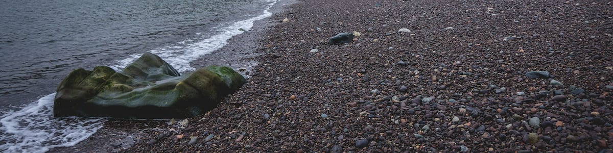 A pebbly beach with waves, a forested cliff, and a rocky outcrop.