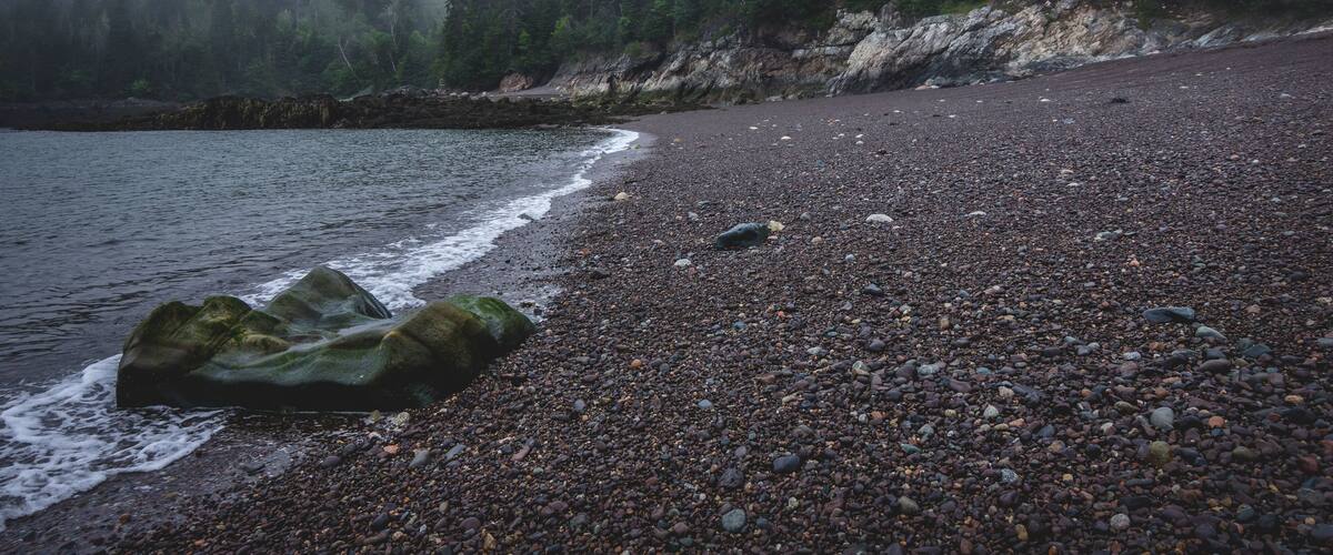 Uma praia de seixos com um afloramento rochoso, águas calmas e um morro coberto de mata.