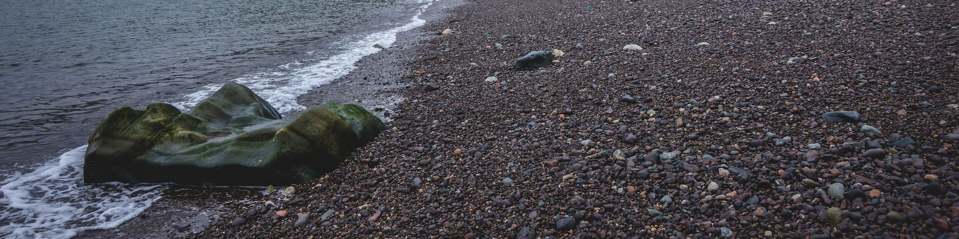 A pebbly beach with a rocky outcrop, calm water, and a forested hillside.