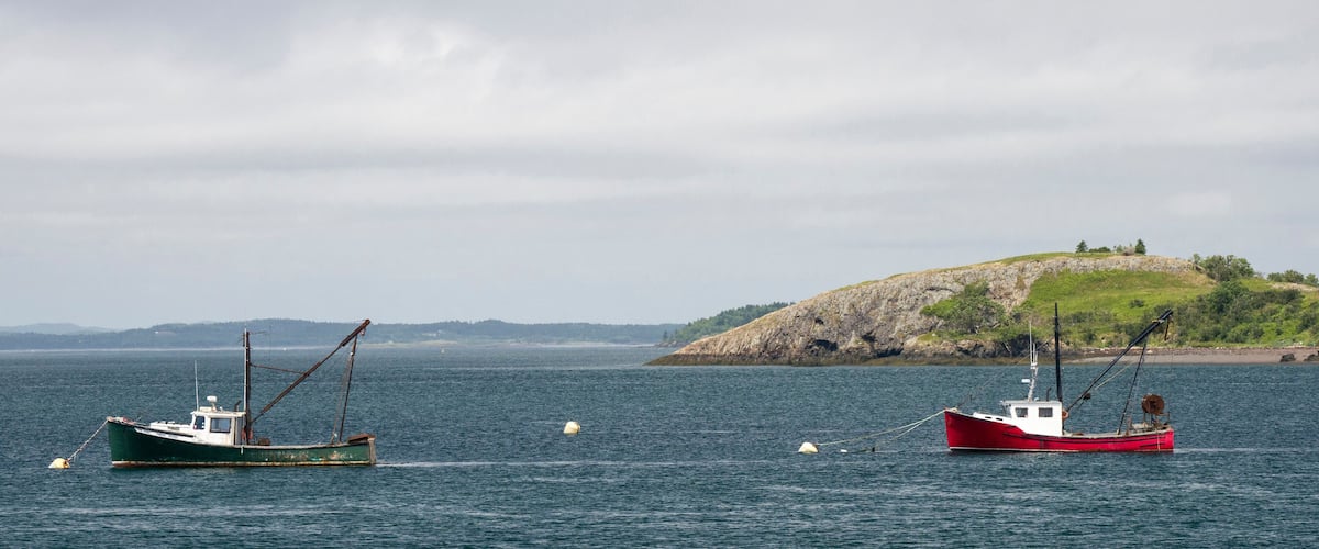 Two fishing boats on calm water with a rocky island and a lighthouse in the background.