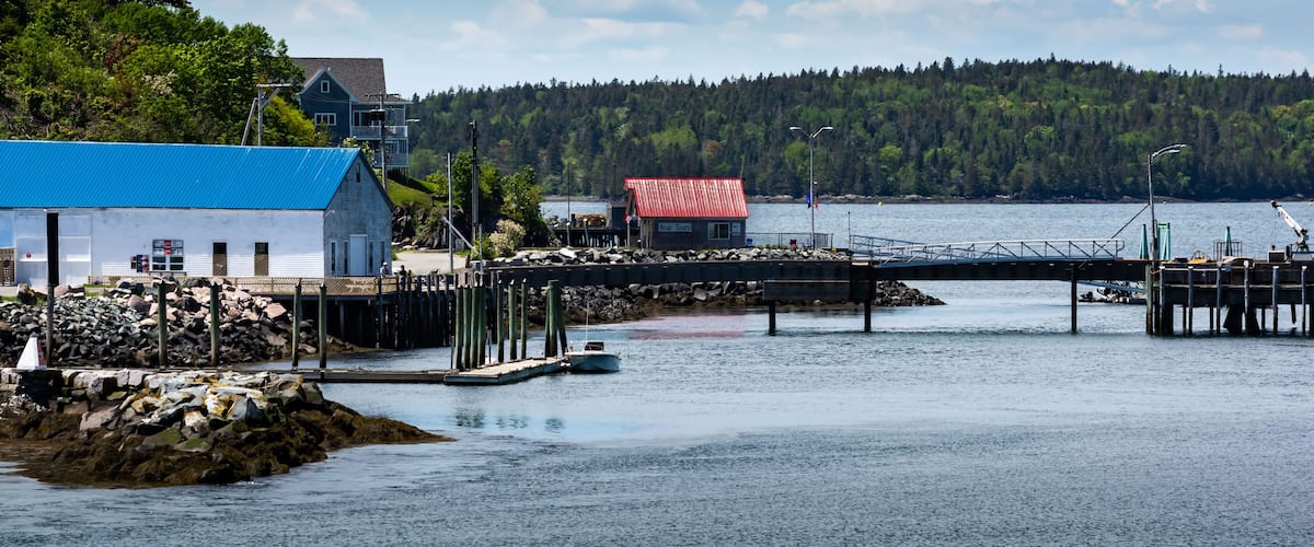 A dock extending into calm waters, a small boat, a building with a blue roof, and a forested hill in the background.