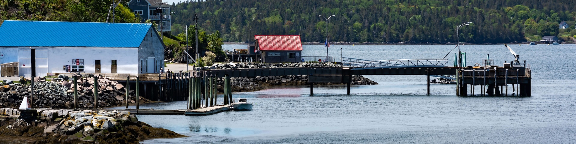 A dock extending into calm waters, a small boat, a building with a blue roof, and a forested hill in the background.