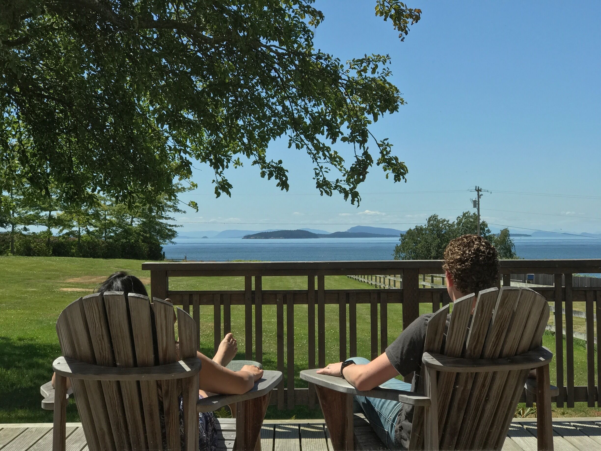 Two people sitting on wooden chairs on a deck overlooking a body of water with distant landforms.