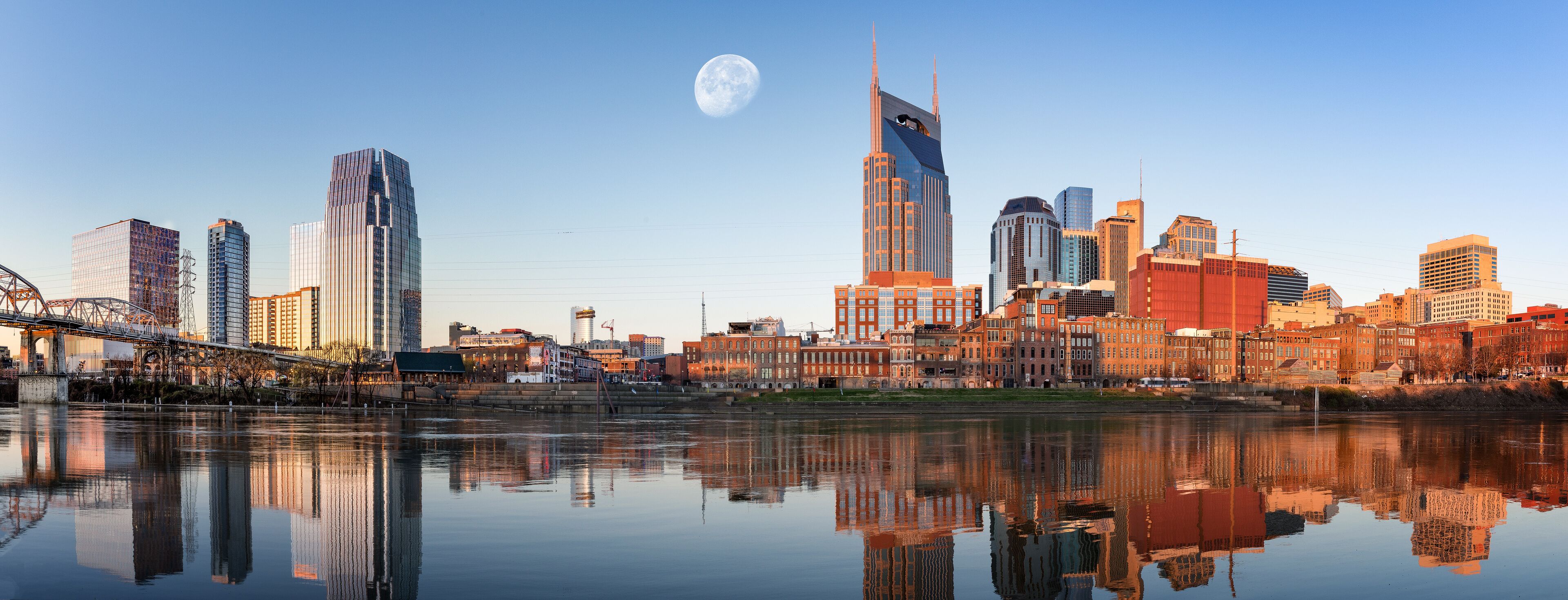 A city skyline with a river reflecting buildings, a bridge, and a full moon in the sky.