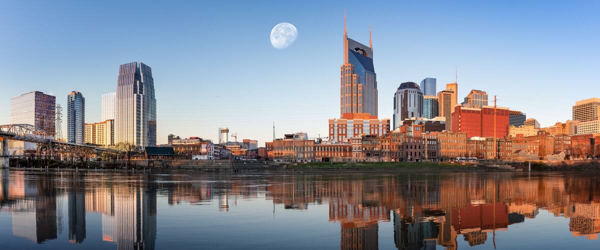 A city skyline with a river reflecting buildings, a bridge, and a full moon in the sky.