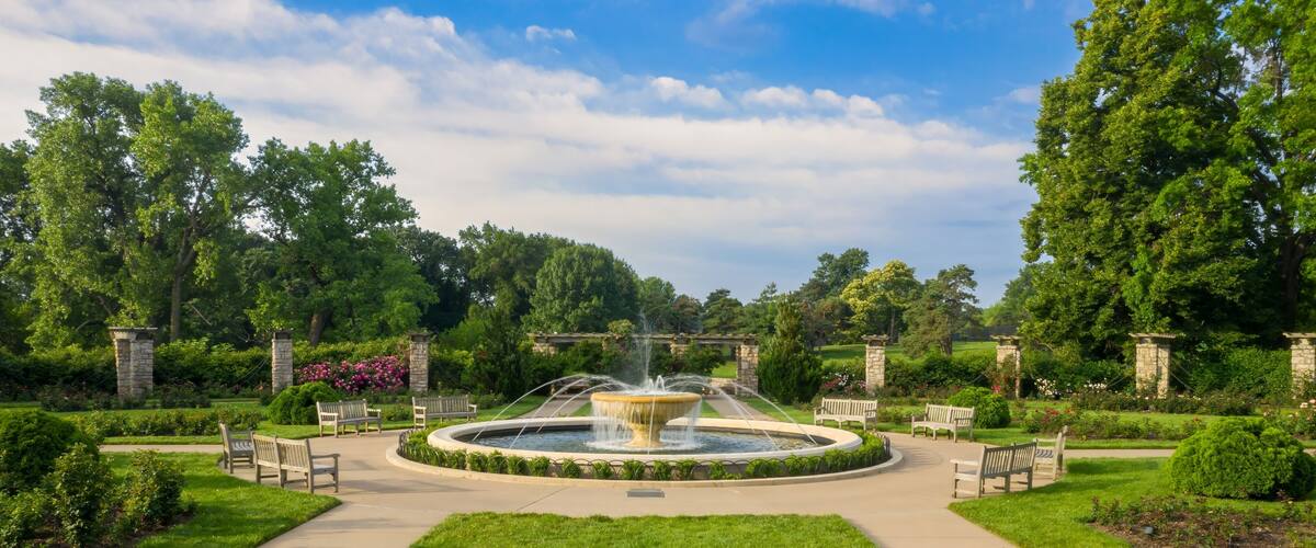 A circular fountain surrounded by a stone pathway, benches, and lush greenery.