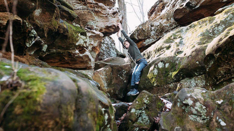 A person climbing a rocky terrain with moss-covered rocks and trees in the background.