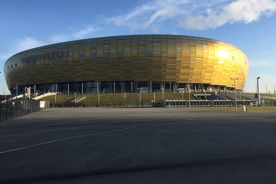 A large, golden stadium with a curved roof and a staircase leading up to the entrance.