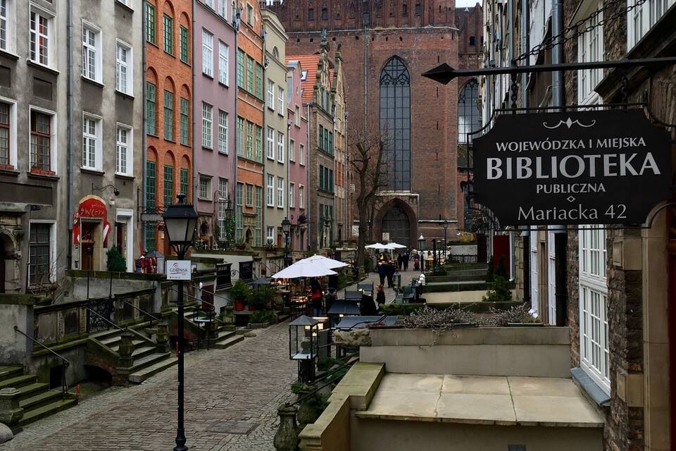 A cobblestone street with colorful buildings, a large church with green spires, and a sign for the Mariacka 42 public library.