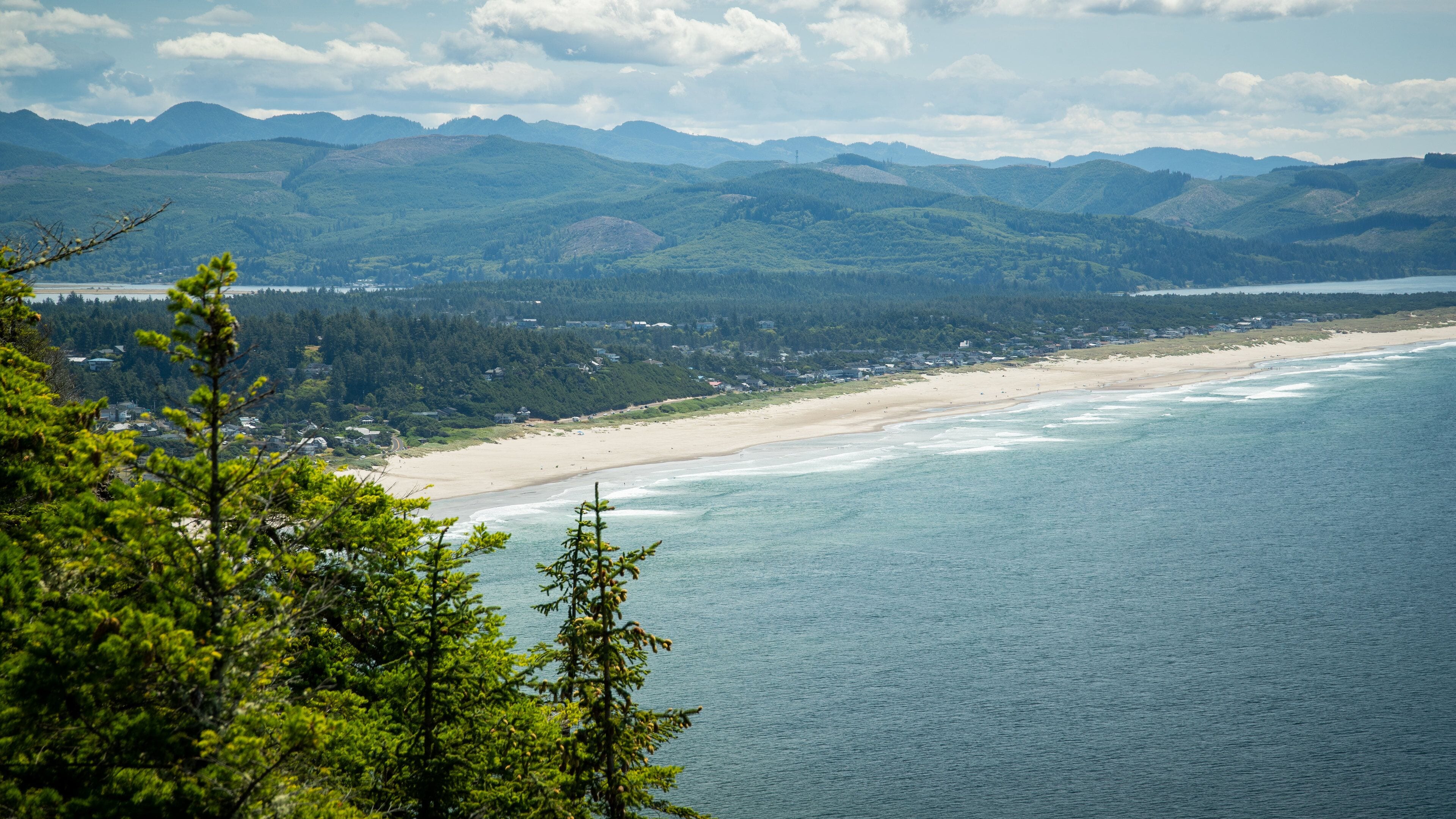 A coastal landscape with a sandy beach, a calm sea, and a backdrop of green hills and mountains.