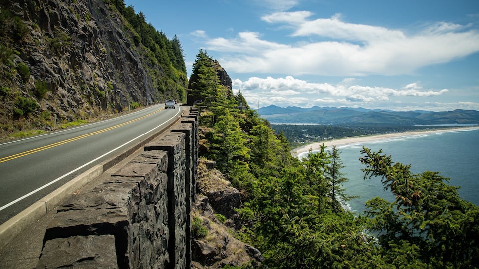 A coastal road with a stone guardrail, a white car, and a beach with mountains in the background.