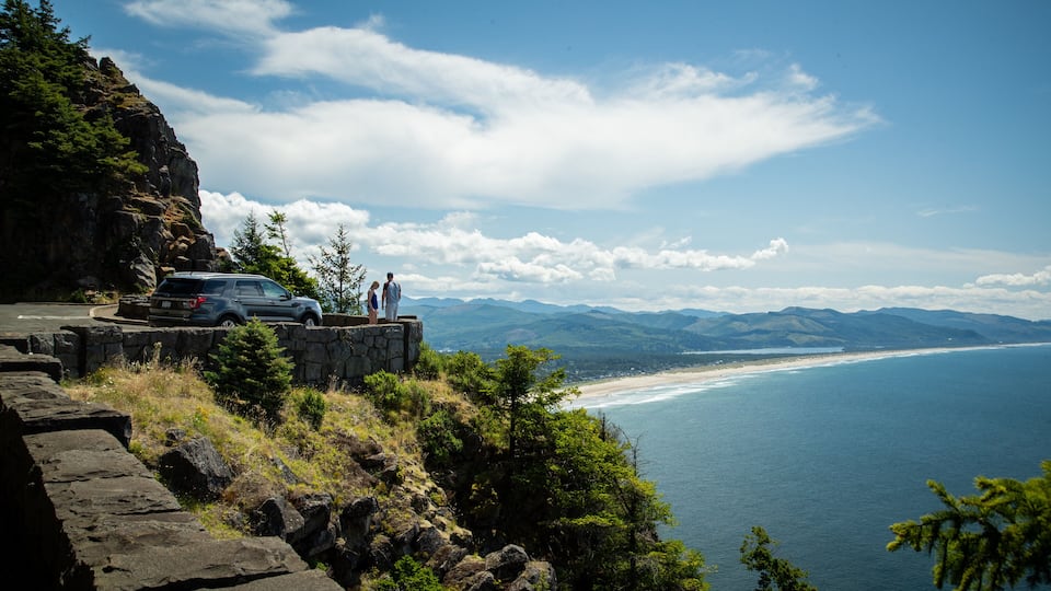 A coastal road with a stone wall, a car, and two people overlooking a beach and mountains.