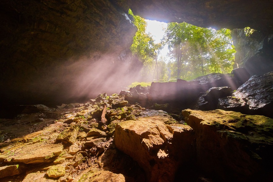 A cave entrance with sunlight streaming through, illuminating the rocks and trees.
