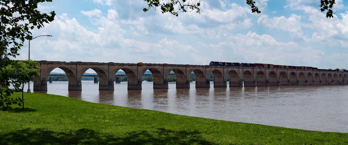 A long bridge spanning a river with multiple arches, a train crossing it, and a grassy bank with trees.