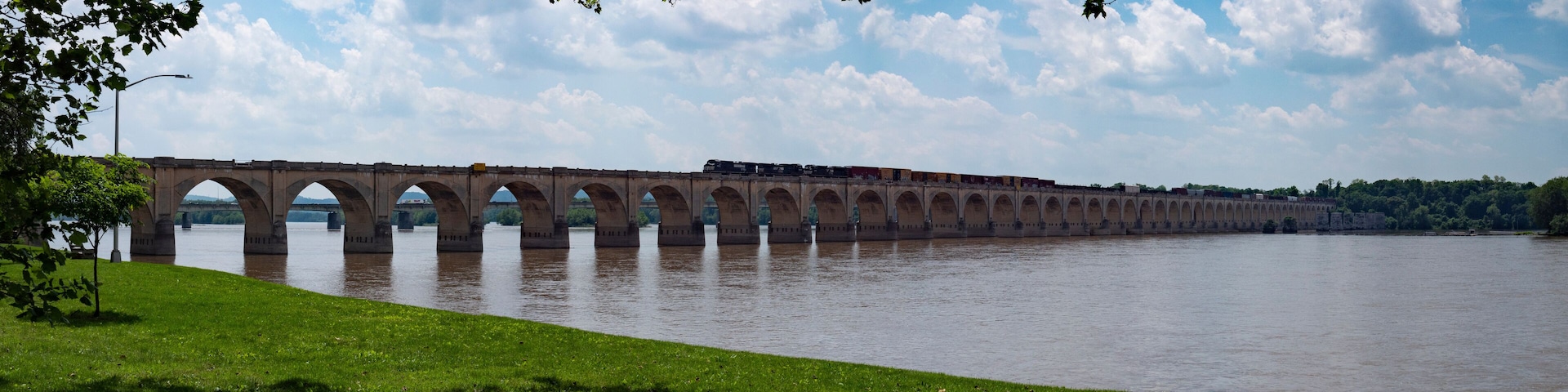 A long bridge spanning a river with multiple arches, a train crossing it, and a grassy bank with trees.