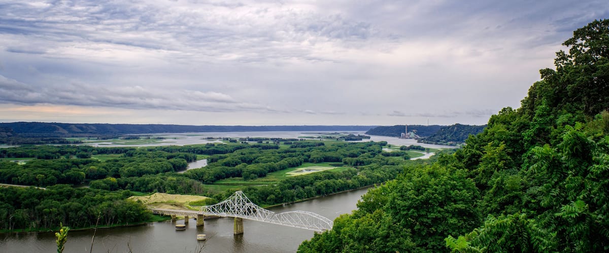 A river with a bridge, surrounded by lush greenery and a cloudy sky.