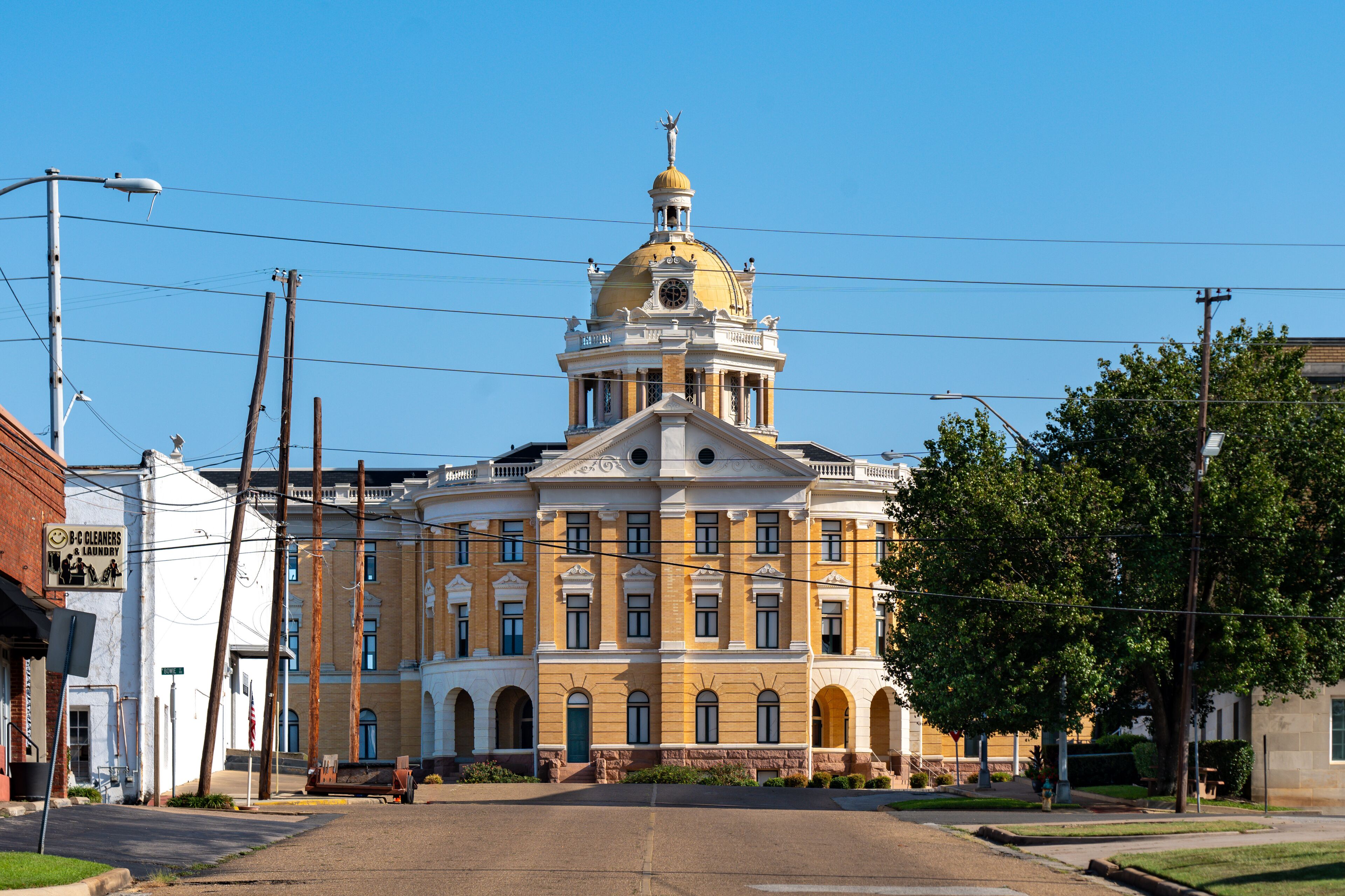 A large yellow building with a dome and columns, surrounded by trees and power lines.