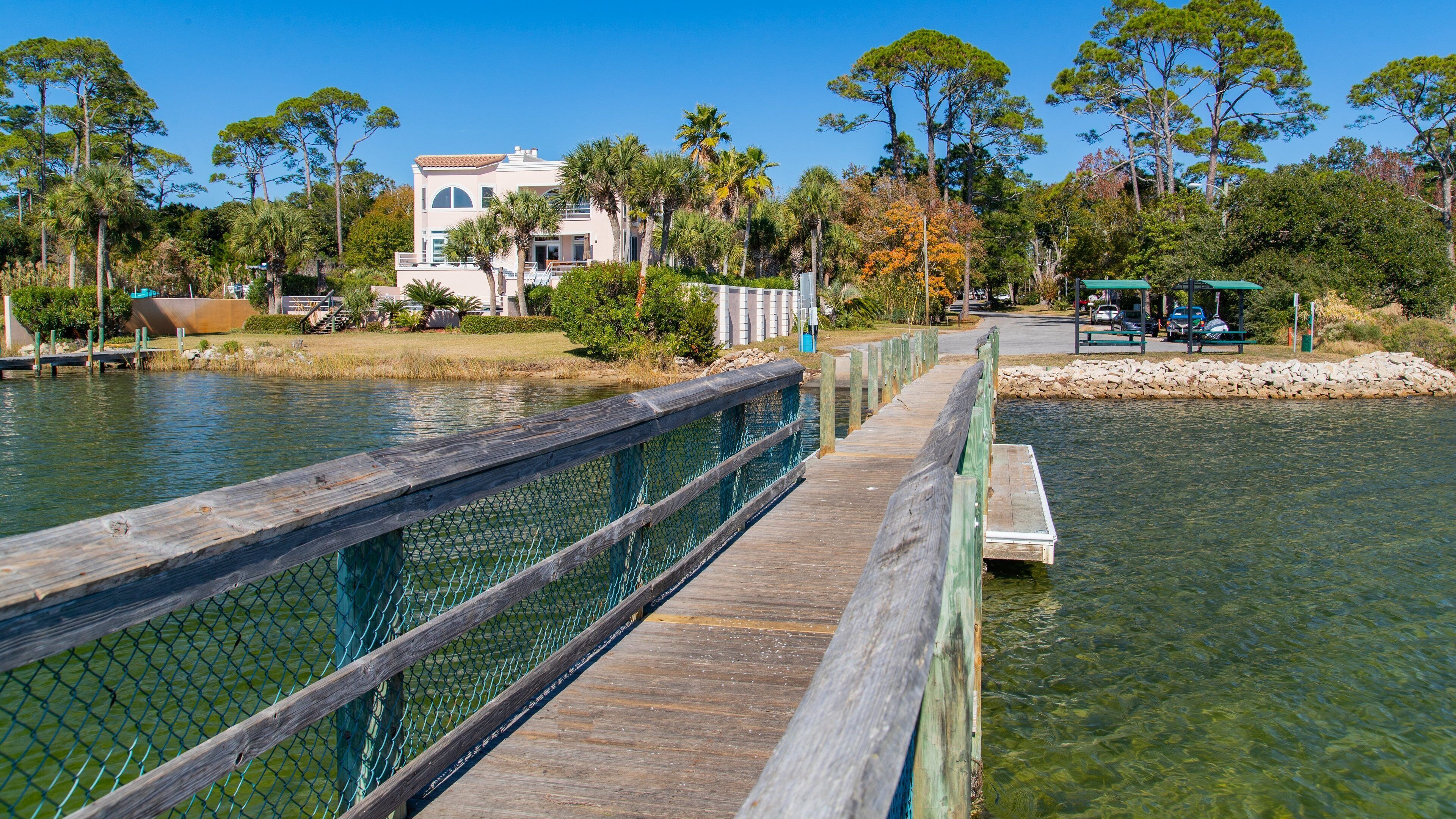 A wooden boardwalk over a calm body of water, surrounded by trees and a building with a white facade.