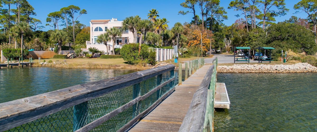 A wooden boardwalk over a calm body of water, surrounded by trees and a building with a white facade.