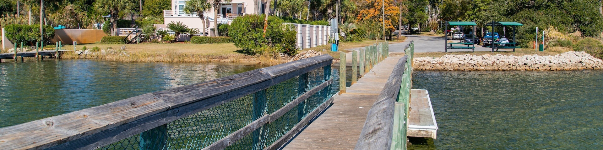 A wooden boardwalk over a calm body of water, surrounded by trees and a building with a white facade.
