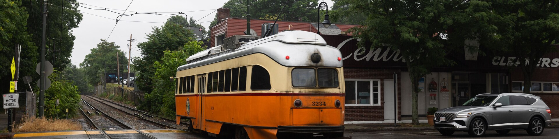 Um elétrico antigo nos trilhos, uma plataforma de estação de trem amarela e branca, e um edifício com uma placa que diz 'CLEANERS'.
