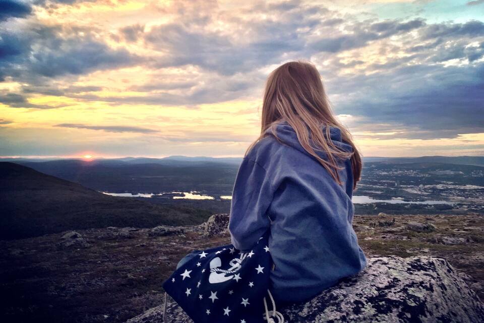 Uma pessoa sentada em uma rocha apreciando um vale com o pôr do sol ao fundo.