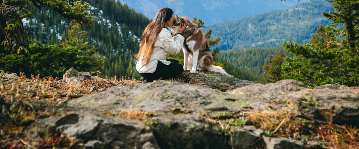Een vrouw en een hond staan op een rotsachtige richel, met een bergketen op de achtergrond.