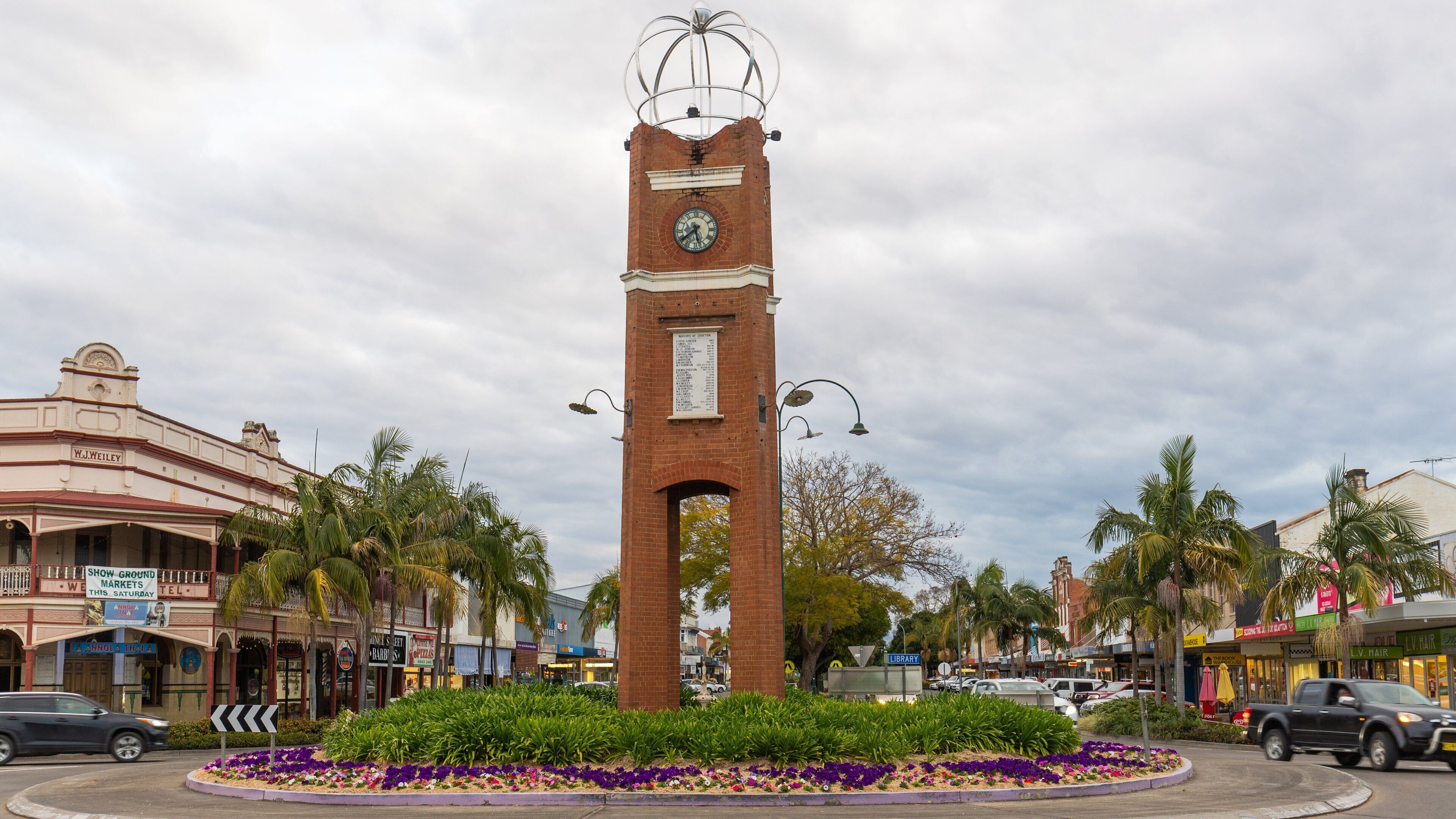 A tall brick clock tower with a clock face, surrounded by a flower bed with purple and white flowers, and palm trees.