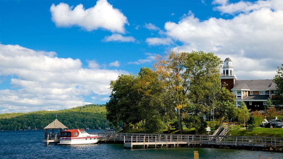 A lake with a boat dock, a gazebo, a church with a bell tower, and a forested hill in the background.