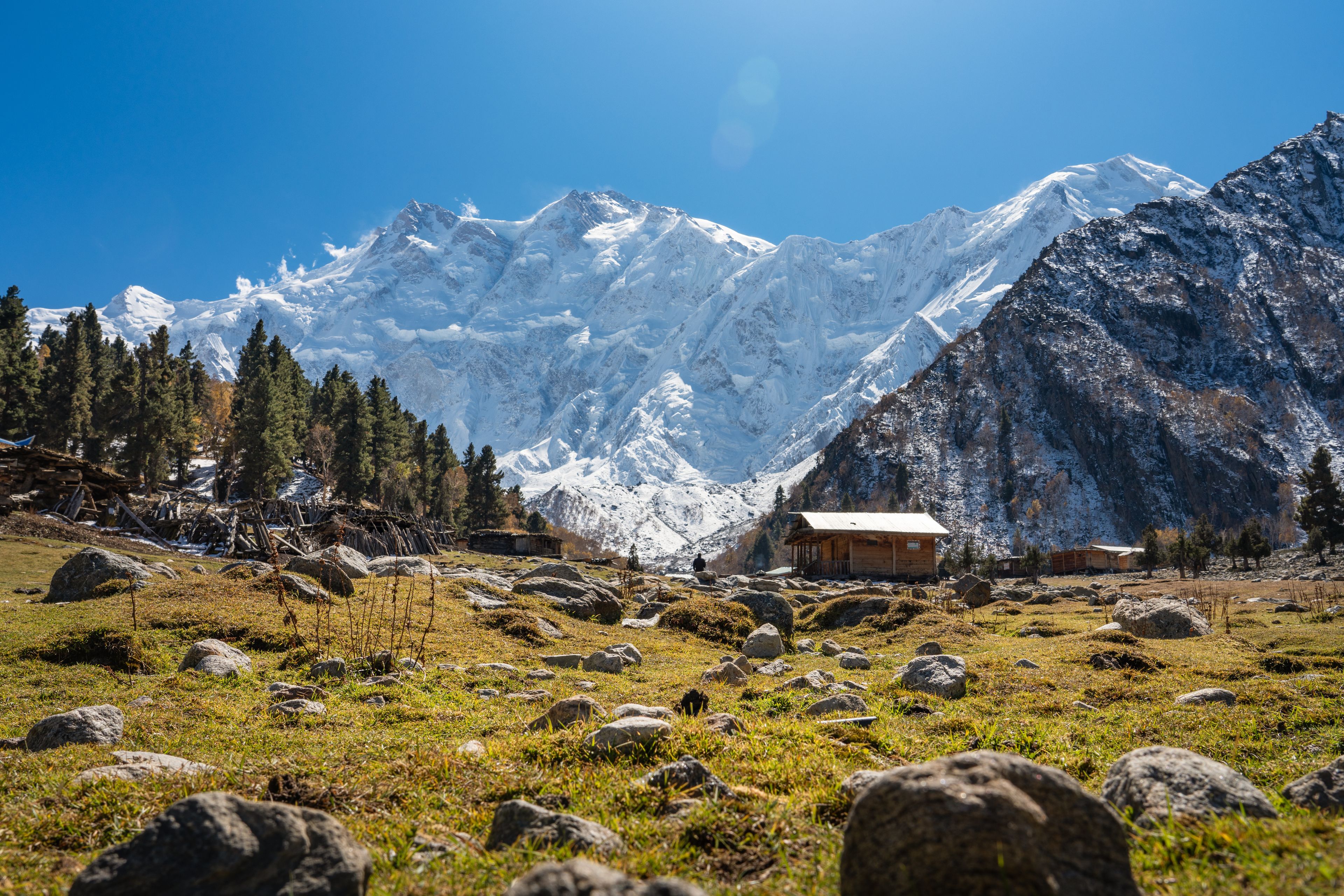 A mountainous landscape with a wooden cabin, rocky terrain, and snow-covered peaks under a clear blue sky.