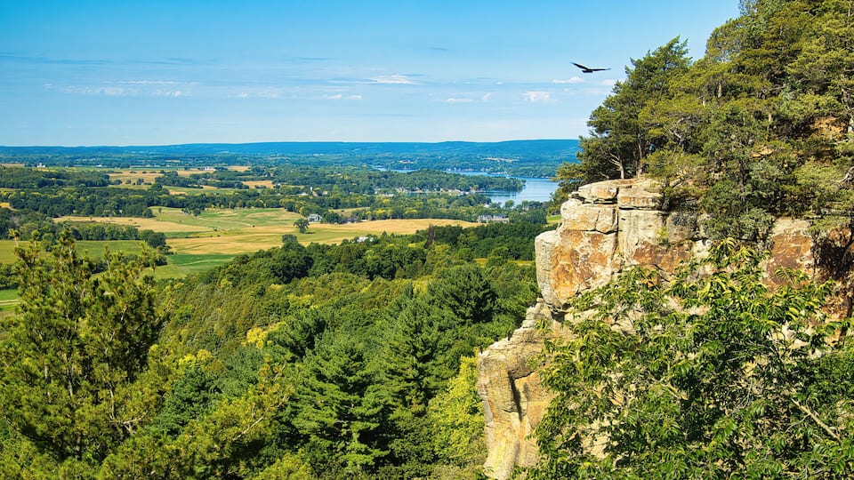 A rocky cliff with dense forest, a river, and distant hills.