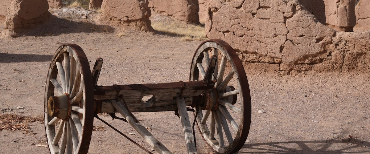 A wooden cart with large wheels in a desert landscape with ancient, earthen structures in the background.