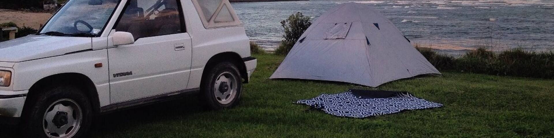 A white car parked on grass next to a tent by a body of water with hills in the background.
