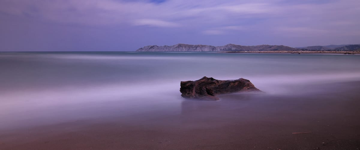 A beach with a rock formation and distant mountains under a purple sky.