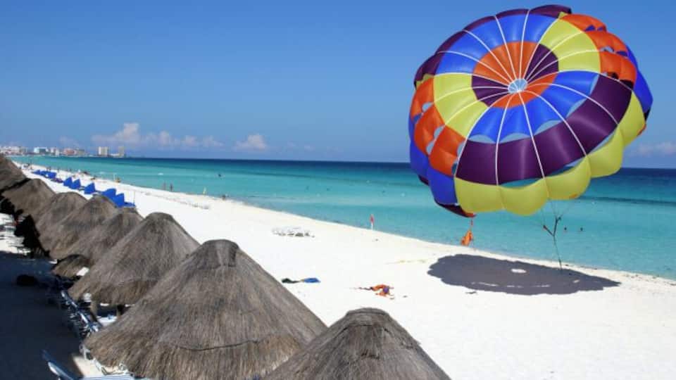 A beach with thatched umbrellas, a colorful parasail, and a clear blue ocean.