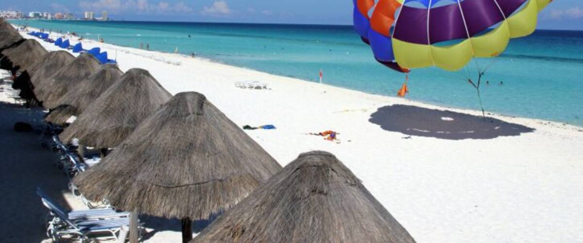 A beach with thatched umbrellas, a colorful parasail, and a clear blue ocean.