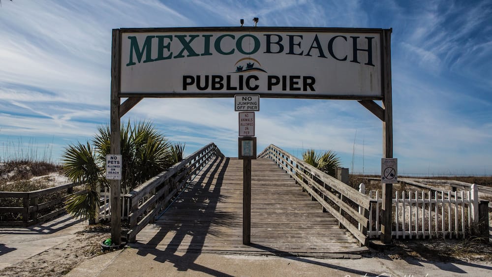 A wooden boardwalk leading to the Mexican Beach Public Pier with signs prohibiting pets and jumping off the pier.