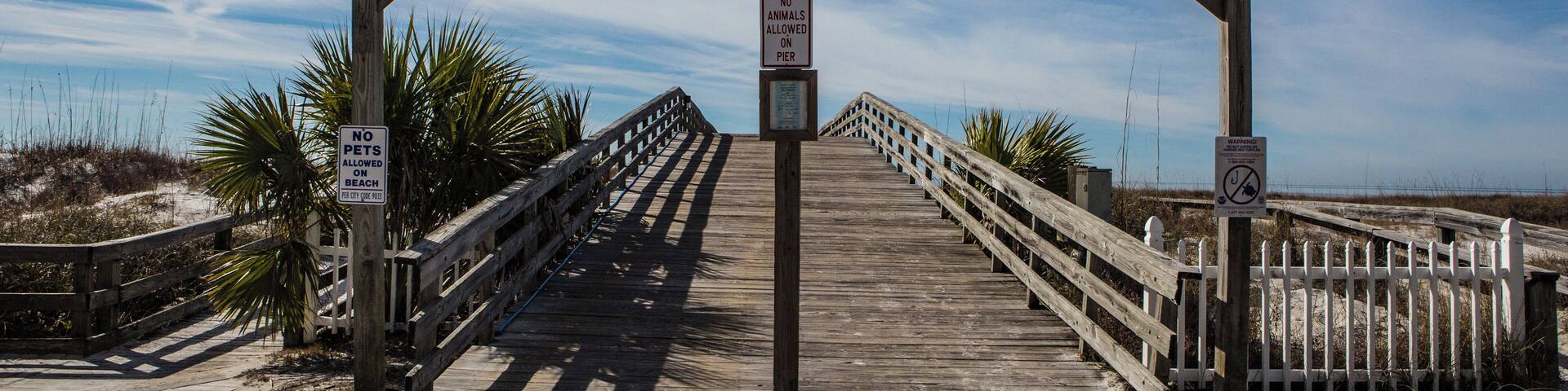 A wooden boardwalk leading to the Mexican Beach Public Pier with signs prohibiting pets and jumping off the pier.