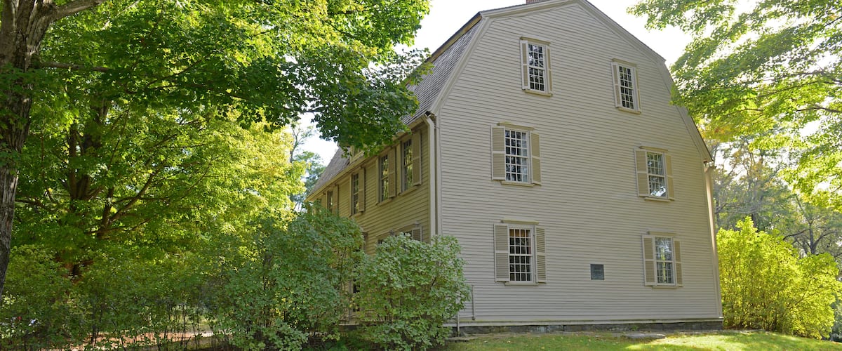 A large, two-story house with a gabled roof, surrounded by lush green trees and a stone wall.