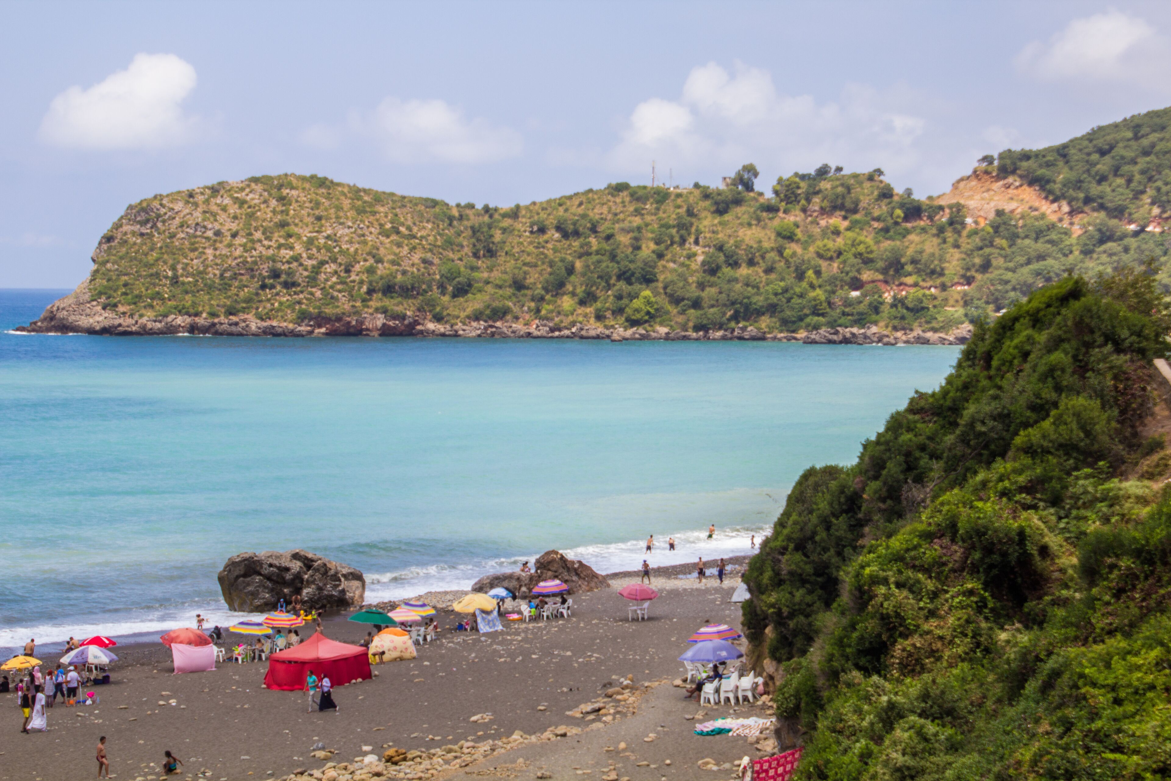 A beach with colorful umbrellas and tents, rocky shoreline, and a hill with dense vegetation in the background.