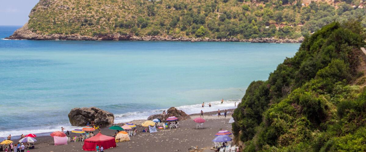 A beach with colorful umbrellas and tents, rocky shoreline, and a hill with dense vegetation in the background.