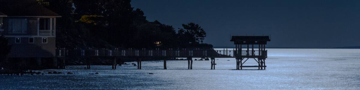 Une nuit illuminée par la lune au-dessus d’une zone riveraine, avec une jetée et un bâtiment.