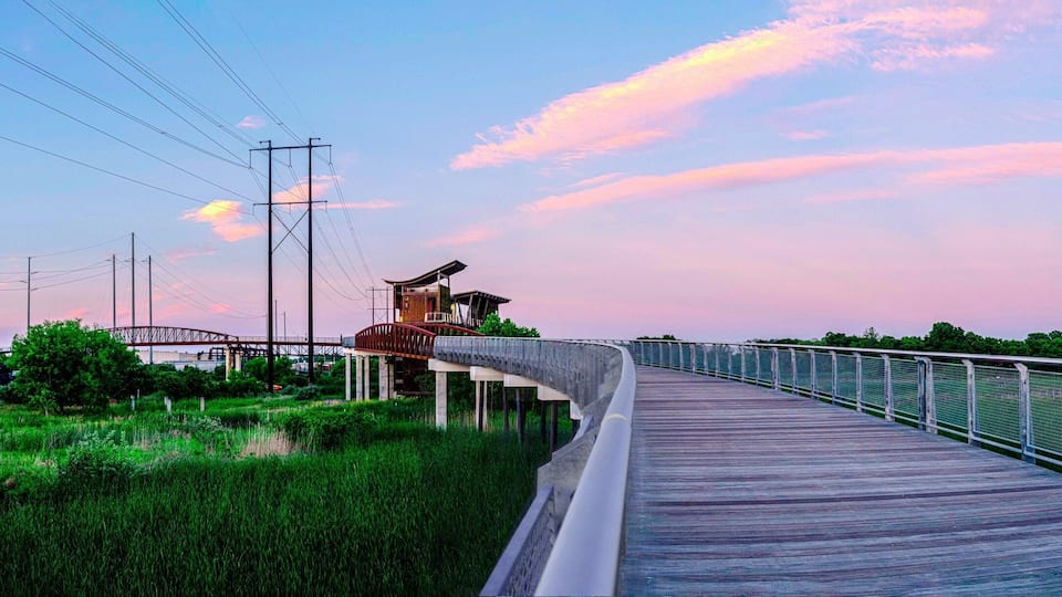 A wooden boardwalk with metal railings, a train track, and a bridge in the background.